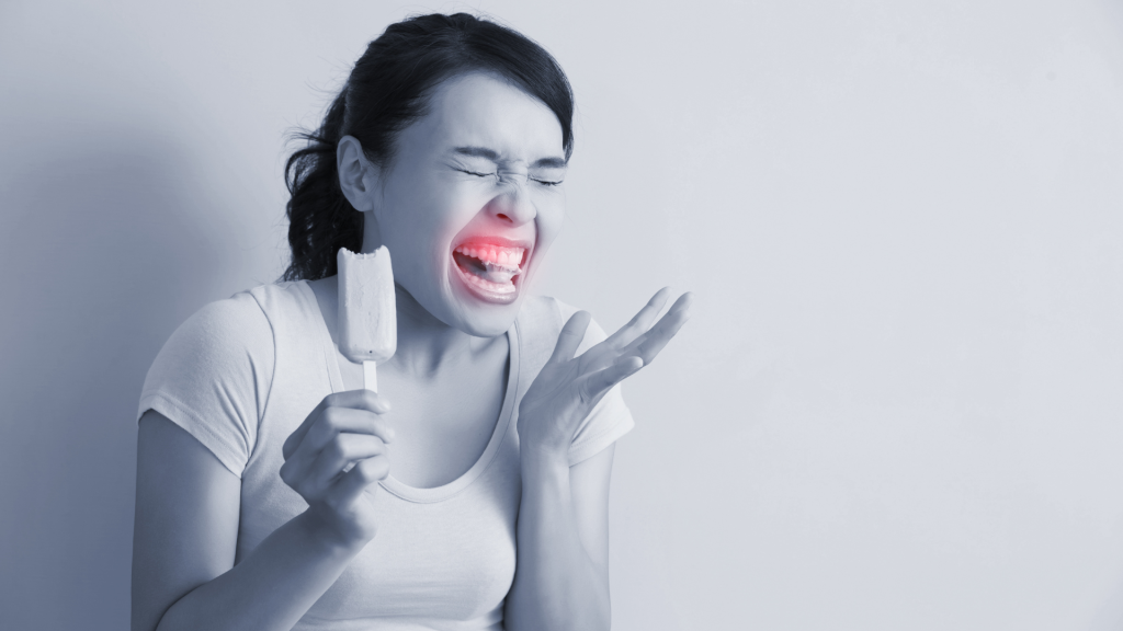 A lady eating an ice lolly, squirming in pain with sensitive teeth. There is a red highlight over her teeth. Image is in black and white.
